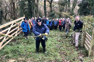 The picture shows His Excellecny, Sir john lorimer, cutting a 'ribbon' of ive across a gateway with trees behind and a group of other people in outdoor gear.