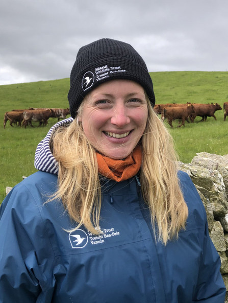 Katie Grace, AES Officer. A young lady smiling at the camera with blond hair wearing a black beanie hat and teal coloured waterproof jacket, both branded with the Manx Wildlife Trust logo